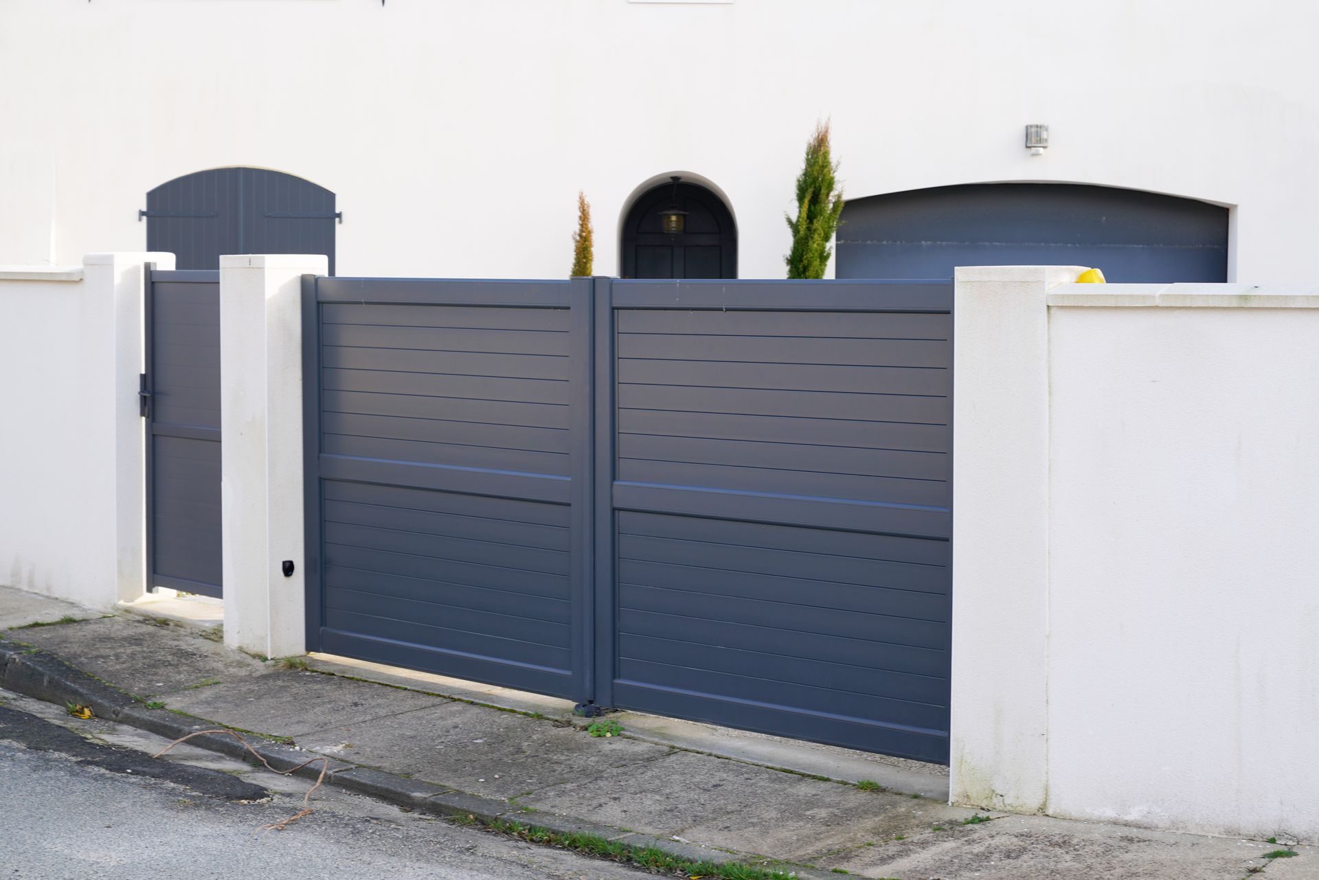 Un portail moderne en métal gris foncé, à lames horizontales, installé entre des piliers en maçonnerie blanche devant une maison.