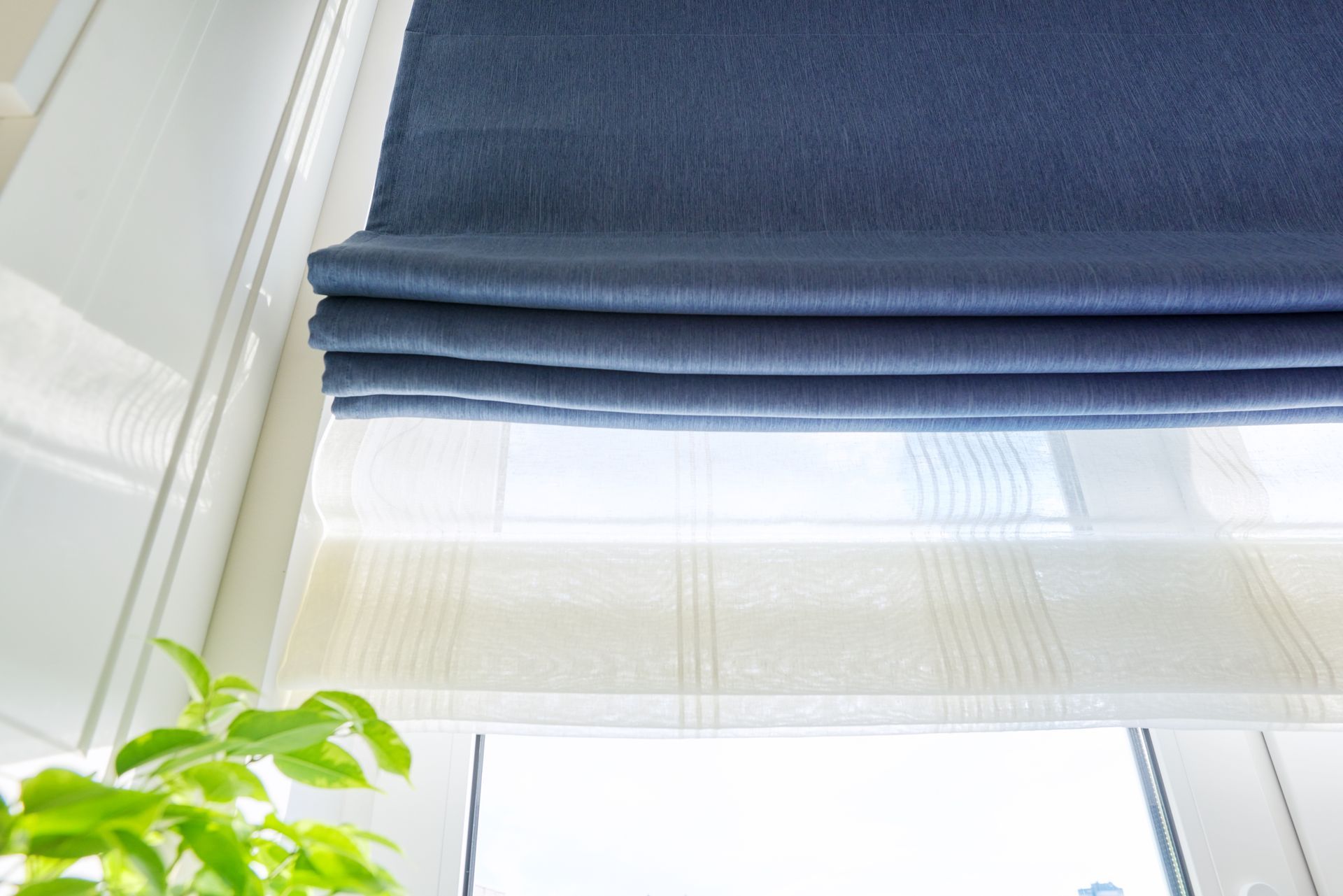 Un store romain en tissu bleu est suspendu au-dessus d'un rideau blanc à rayures transparentes, dans une fenêtre lumineuse avec une plante verte dans le coin.