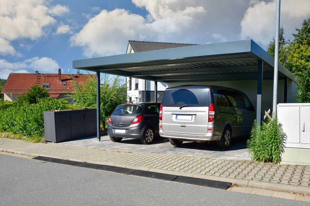 Deux voitures garées sous un abri métallique moderne à côté d'une maison par une journée ensoleillée.