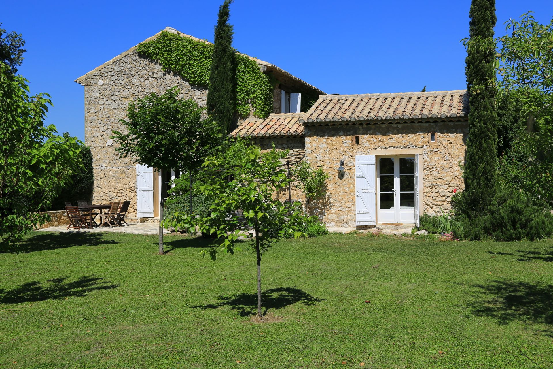 Un cottage en pierre aux volets blancs et recouvert de lierre se dresse au milieu d'un jardin verdoyant, sous un ciel d'un bleu limpide.