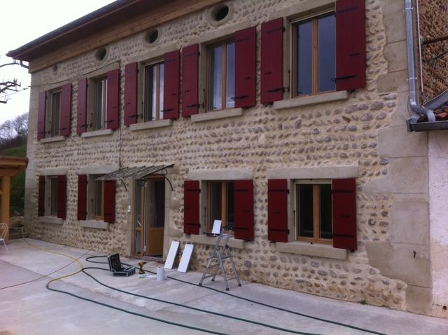 Une ferme en pierre aux volets rouges donnant sur un patio, en cours de rénovation, avec des outils et des matériaux jonchant le sol.