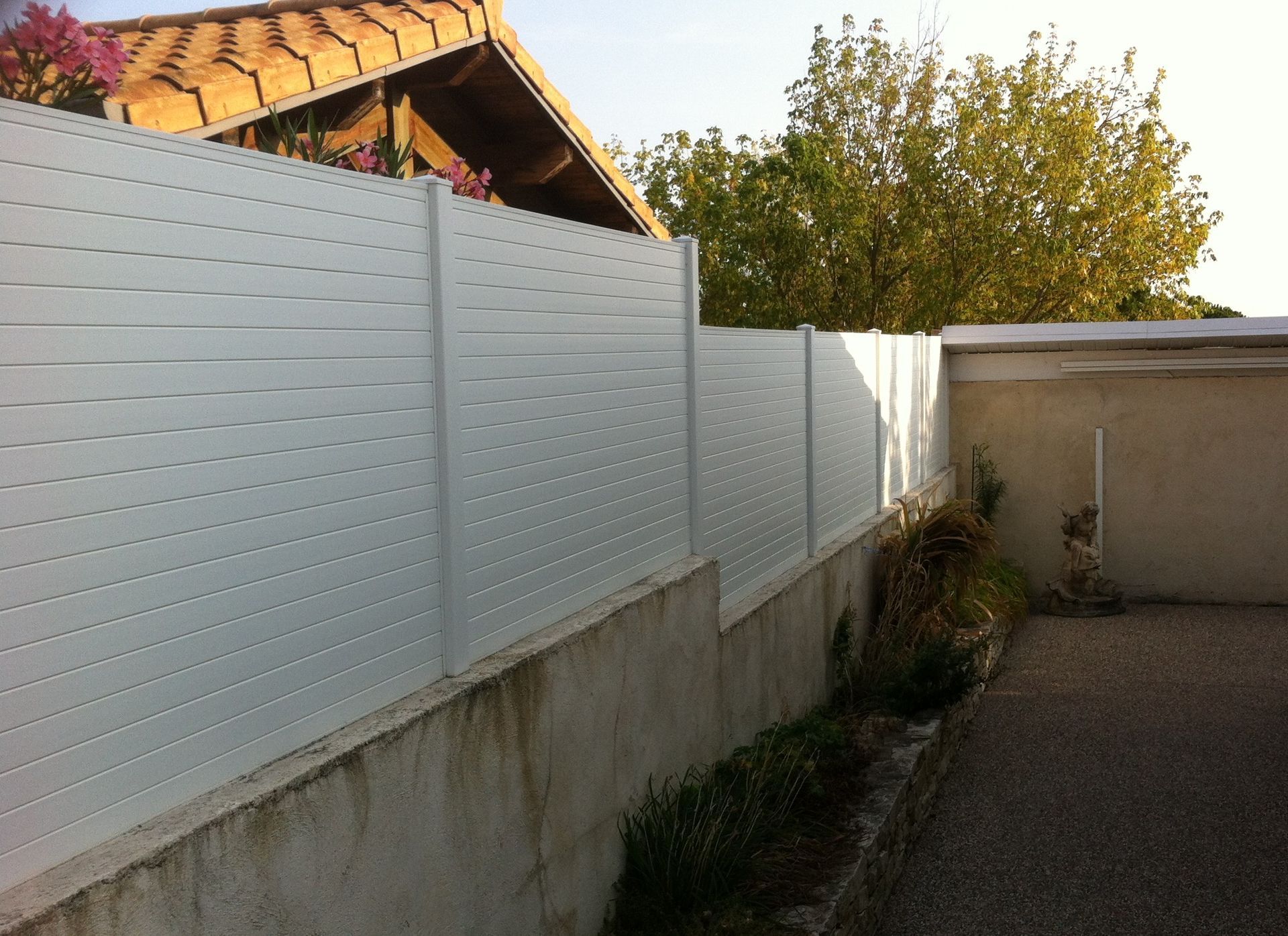 Une clôture métallique blanche à lattes horizontales, fixée sur un mur en béton à côté d'un chemin de gravier, assure l'intimité.
