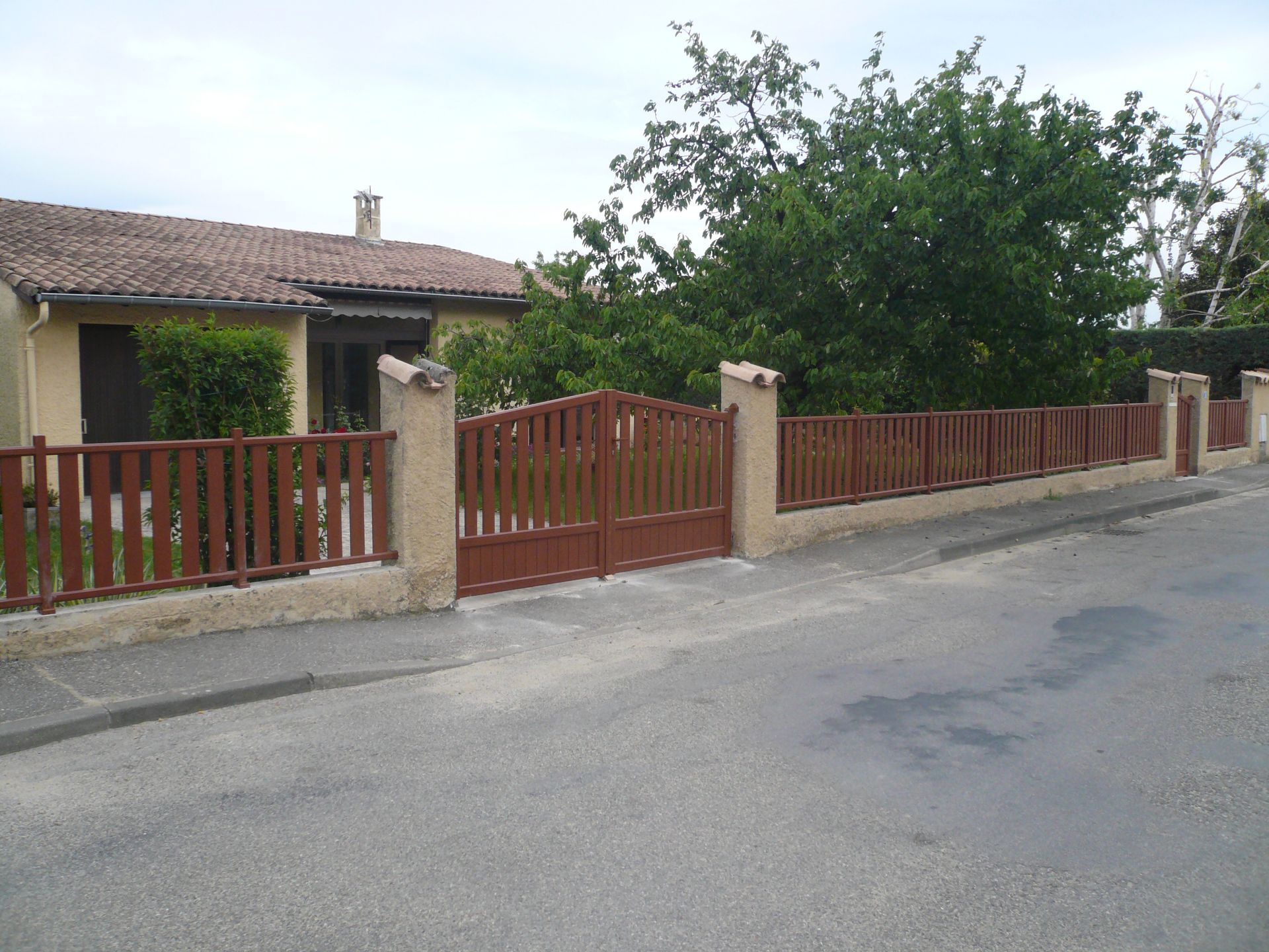 Une maison de plain-pied au toit de tuiles, derrière une clôture et un portail en bois brun, située au bord d'une route pavée.