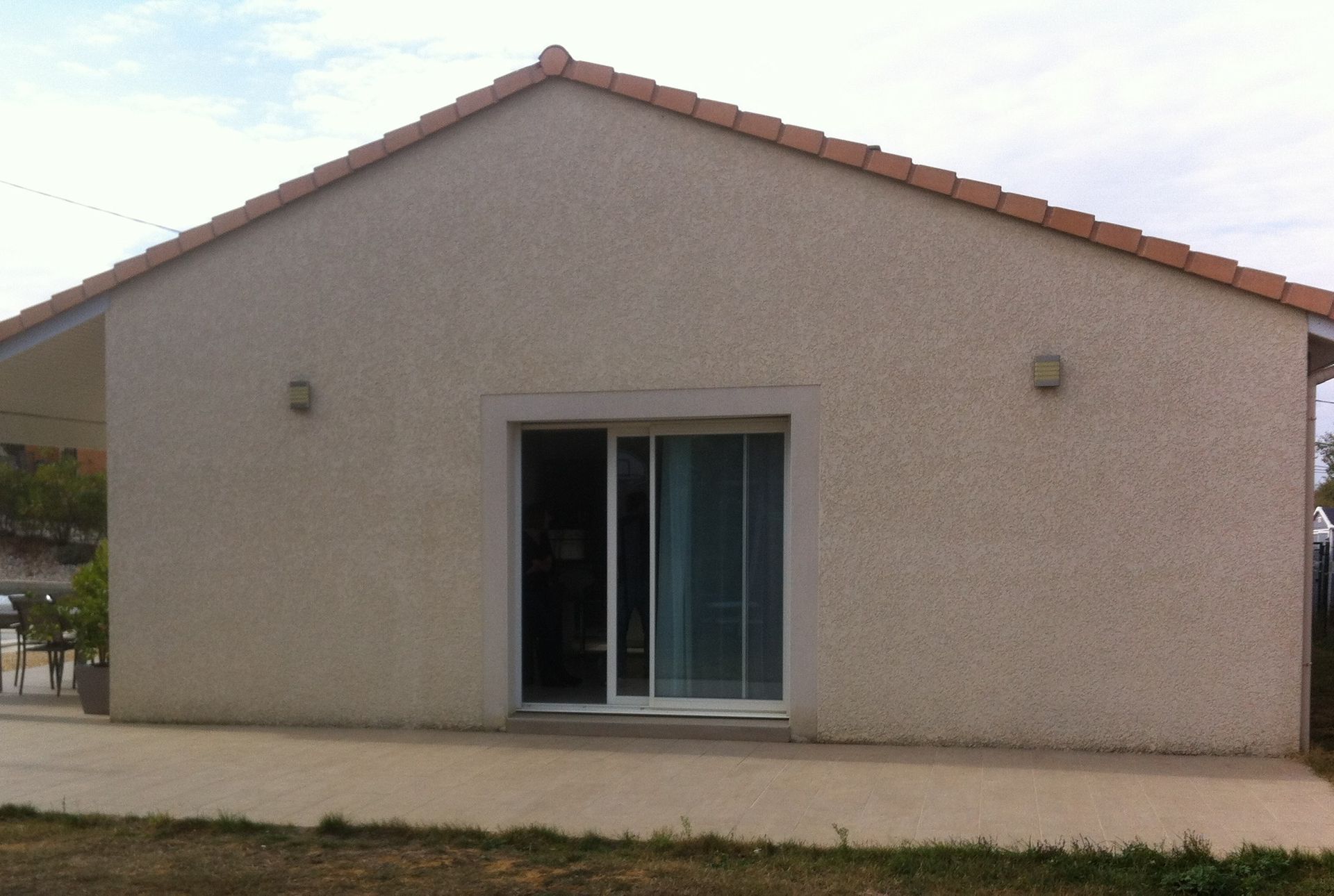 Maison beige de plain-pied avec un toit à pignon, une porte-fenêtre coulissante centrale et une terrasse en béton attenante.