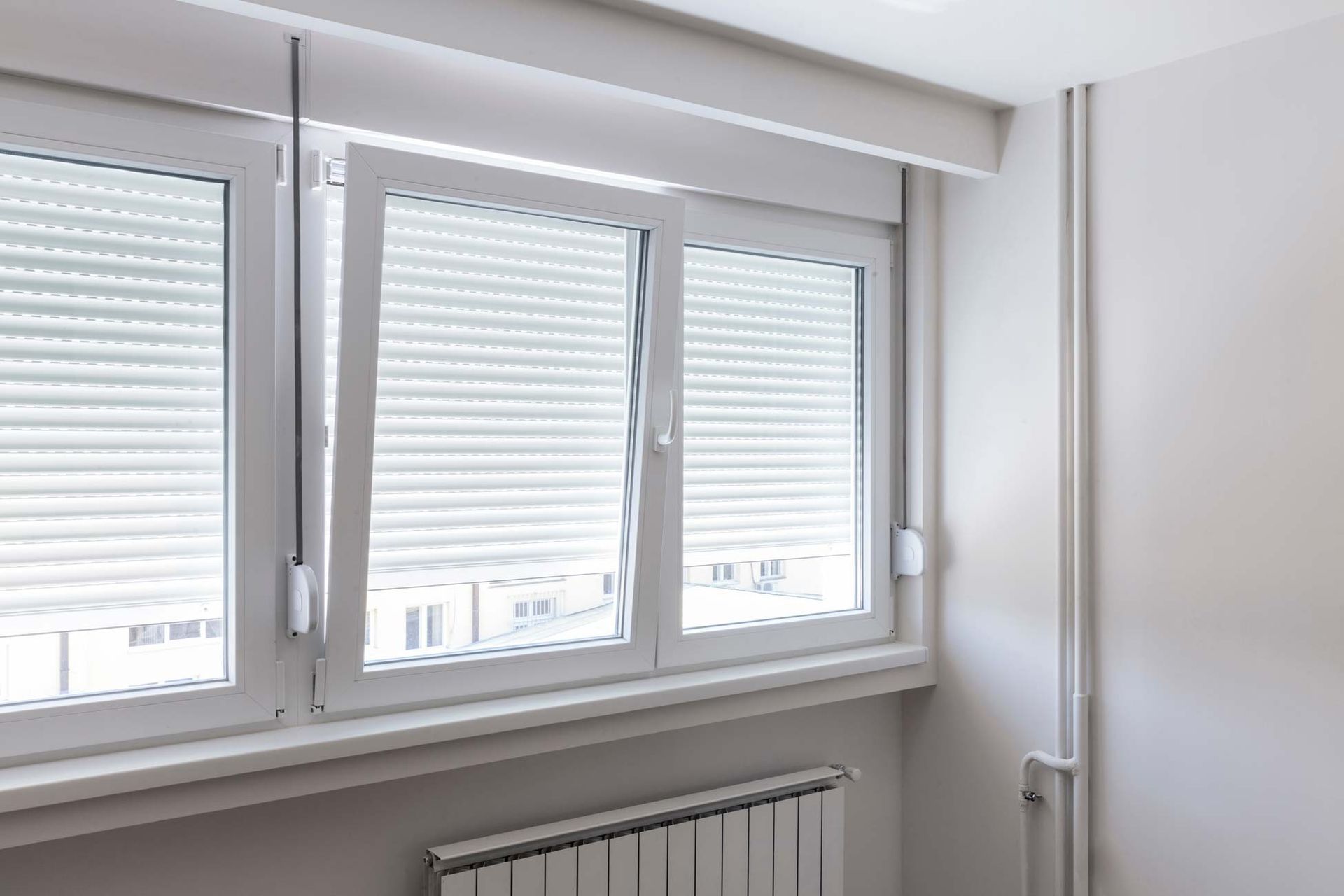 Une fenêtre blanche à plusieurs carreaux, avec des volets roulants abaissés, placée au-dessus d'un radiateur dans une pièce minimaliste.