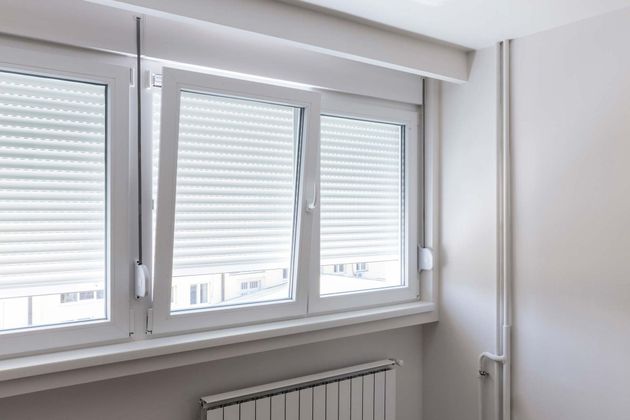 Une fenêtre blanche à plusieurs carreaux, avec des volets roulants abaissés, placée au-dessus d'un radiateur dans une pièce minimaliste.