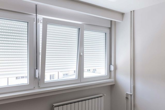 Une fenêtre blanche à plusieurs carreaux, avec des volets roulants abaissés, placée au-dessus d'un radiateur dans une pièce minimaliste.