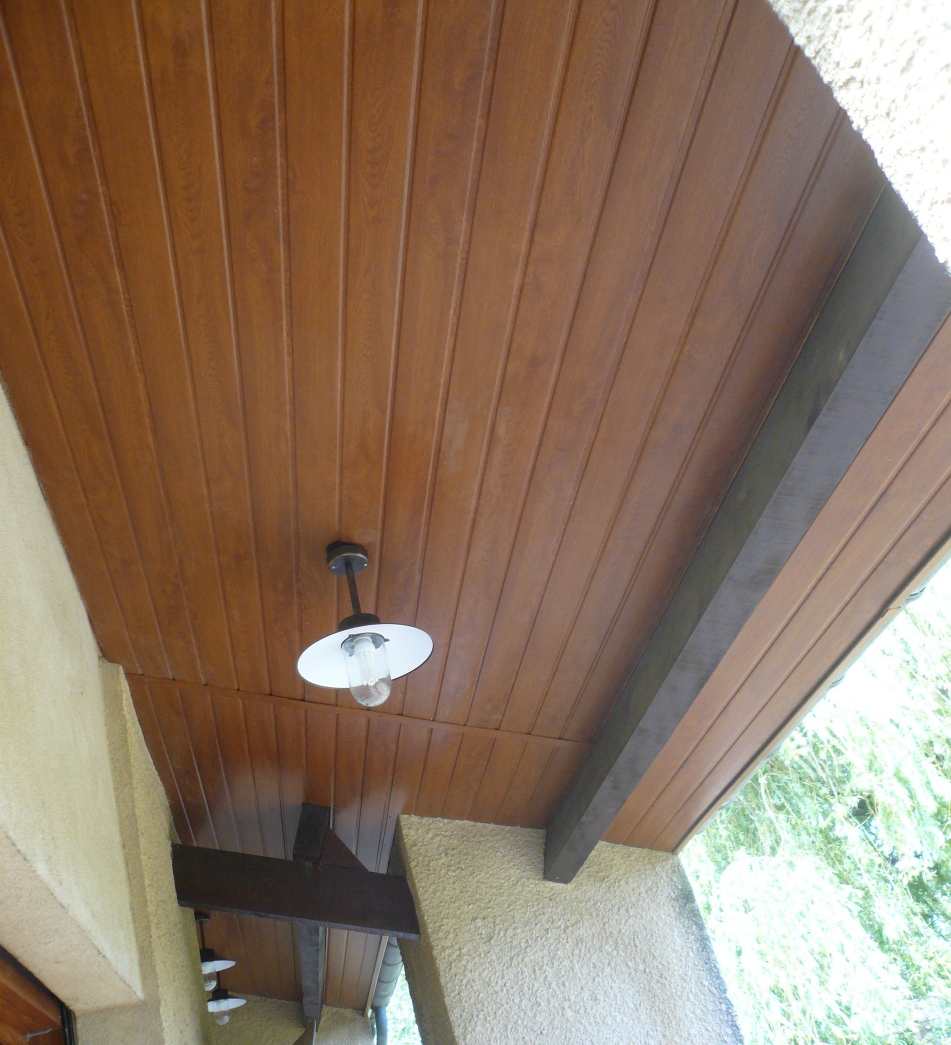 Un plafond de véranda lambrissé avec une suspension lumineuse et des poutres de soutien sombres contre un mur en stuc.