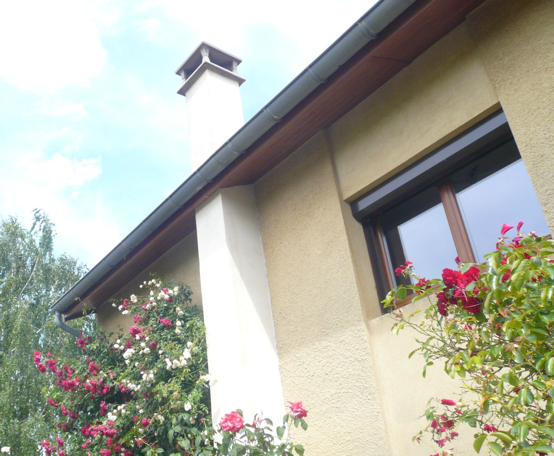 Vue en contreplongée de l'extérieur d'une maison beige avec une cheminée blanche, un encadrement de fenêtre marron et des roses roses et blanches en fleurs.