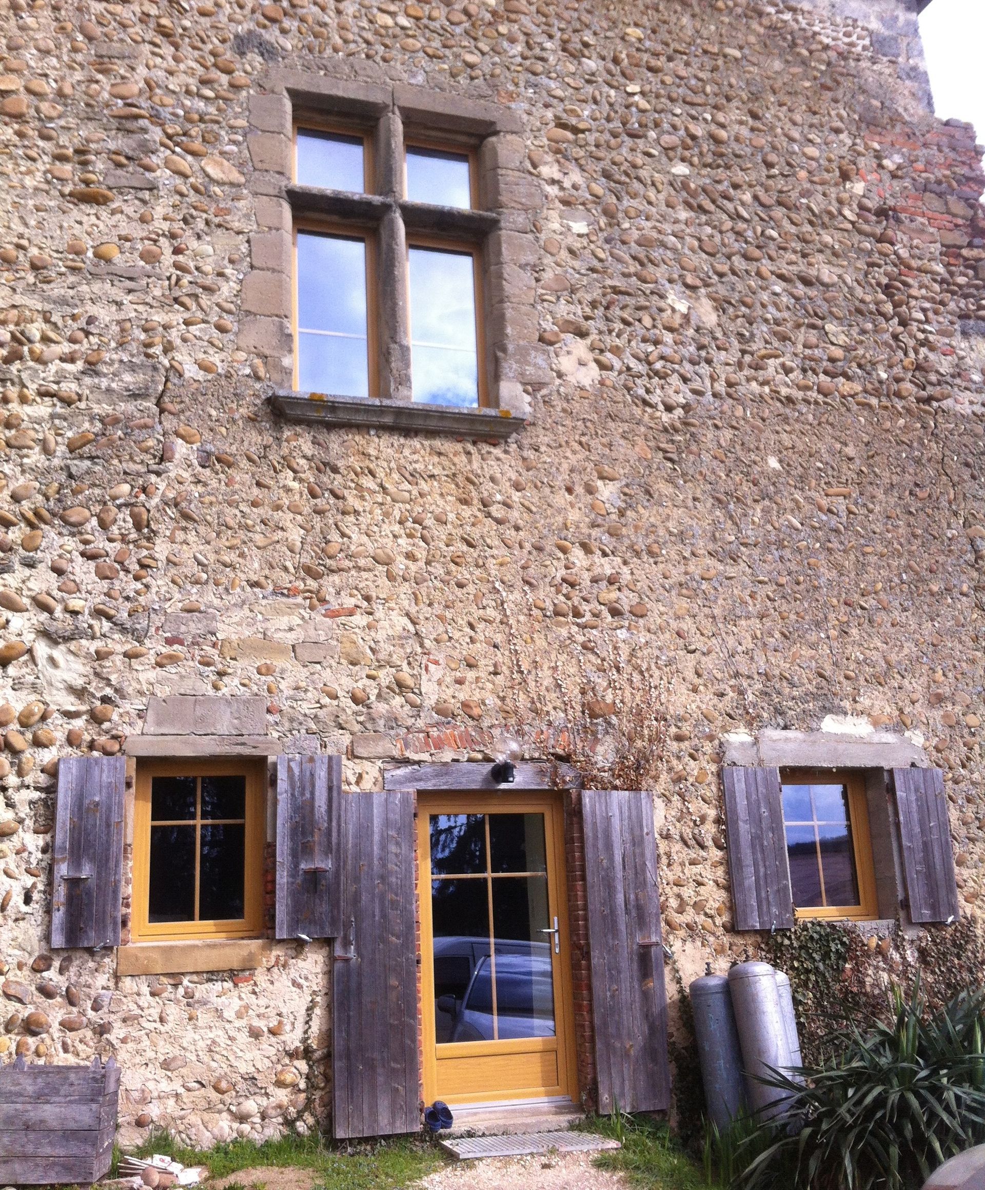 L'extérieur rustique d'une maison en pierre présente une grande fenêtre à l'étage, une porte centrale en bois et deux fenêtres latérales avec volets.