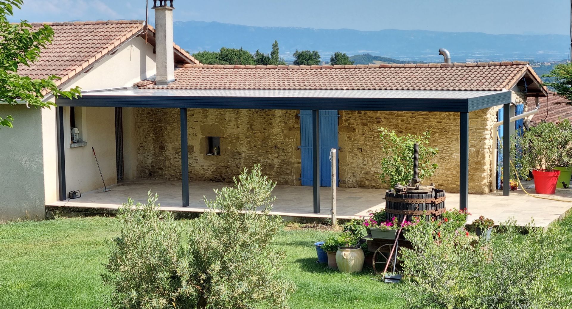 Une maison en pierre avec une terrasse moderne couverte en métal à toit plat, une pelouse verdoyante et des montagnes au loin sous un ciel bleu.