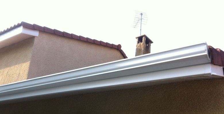 Vue en contreplongée d'une gouttière horizontale blanche sur un mur en stuc beige, avec un toit et une cheminée visibles au-dessus.