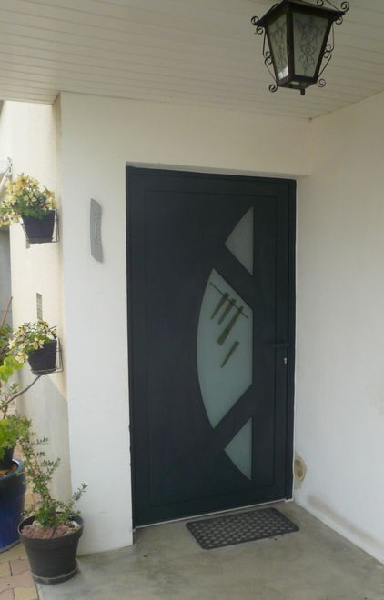 Une porte extérieure gris foncé avec une fenêtre en verre dépoli en forme de croissant, flanquée de plantes en pot sur un porche blanc.