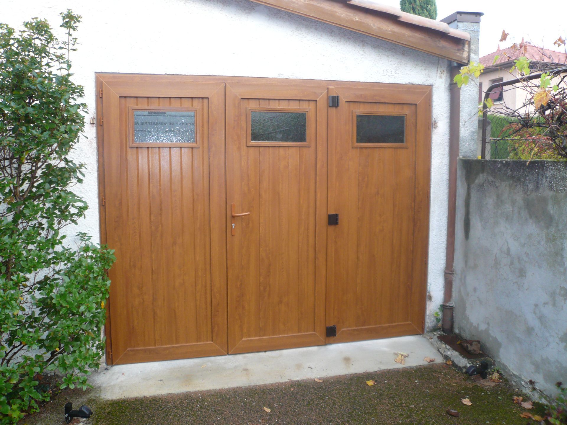 Une porte de garage en bois brun à trois panneaux et petites fenêtres rectangulaires, encastrée dans un mur extérieur blanc.