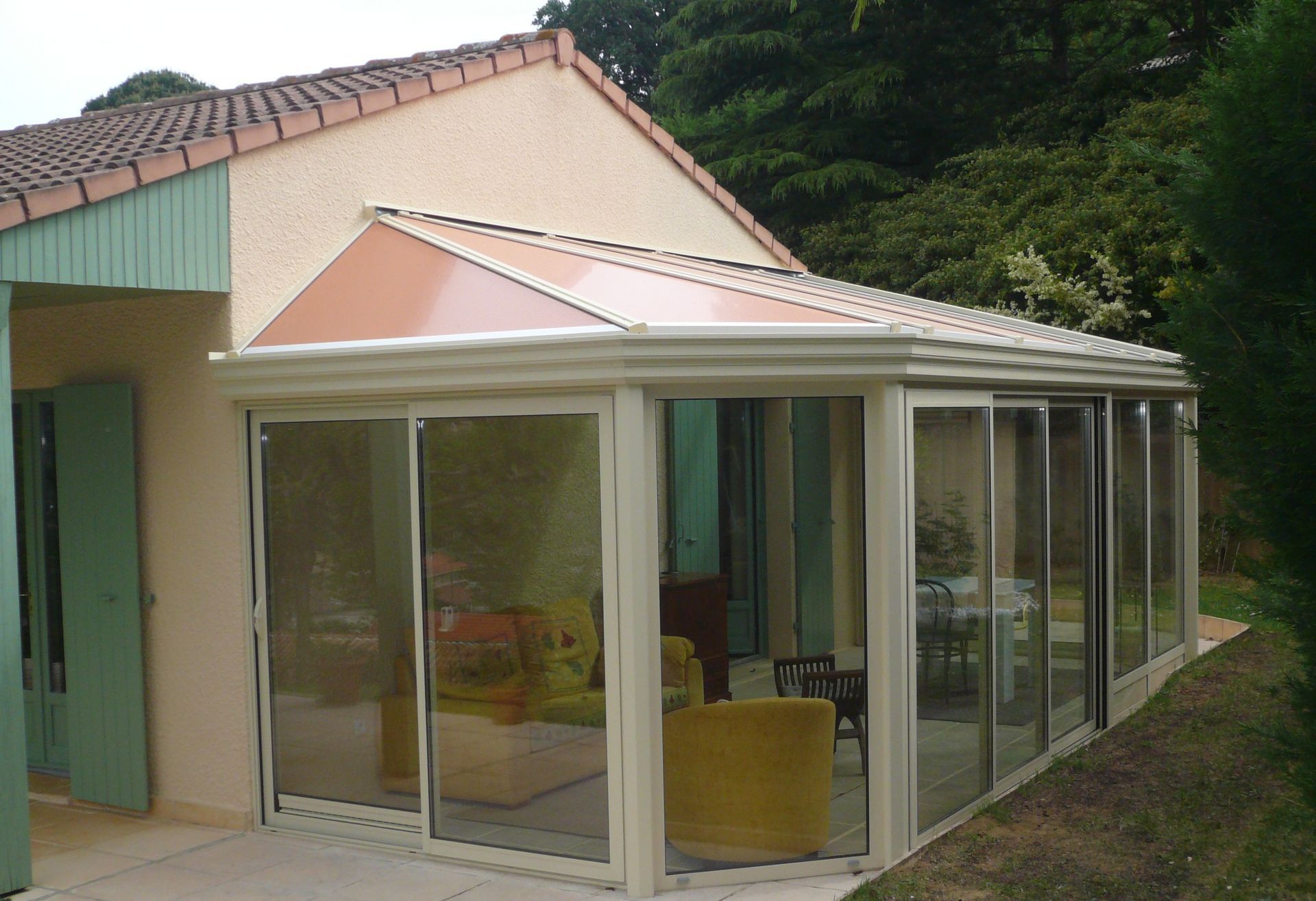 Une maison beige avec une véranda moderne aux parois de verre, dotée d'un toit de tuiles et de portes coulissantes, située dans un jardin.