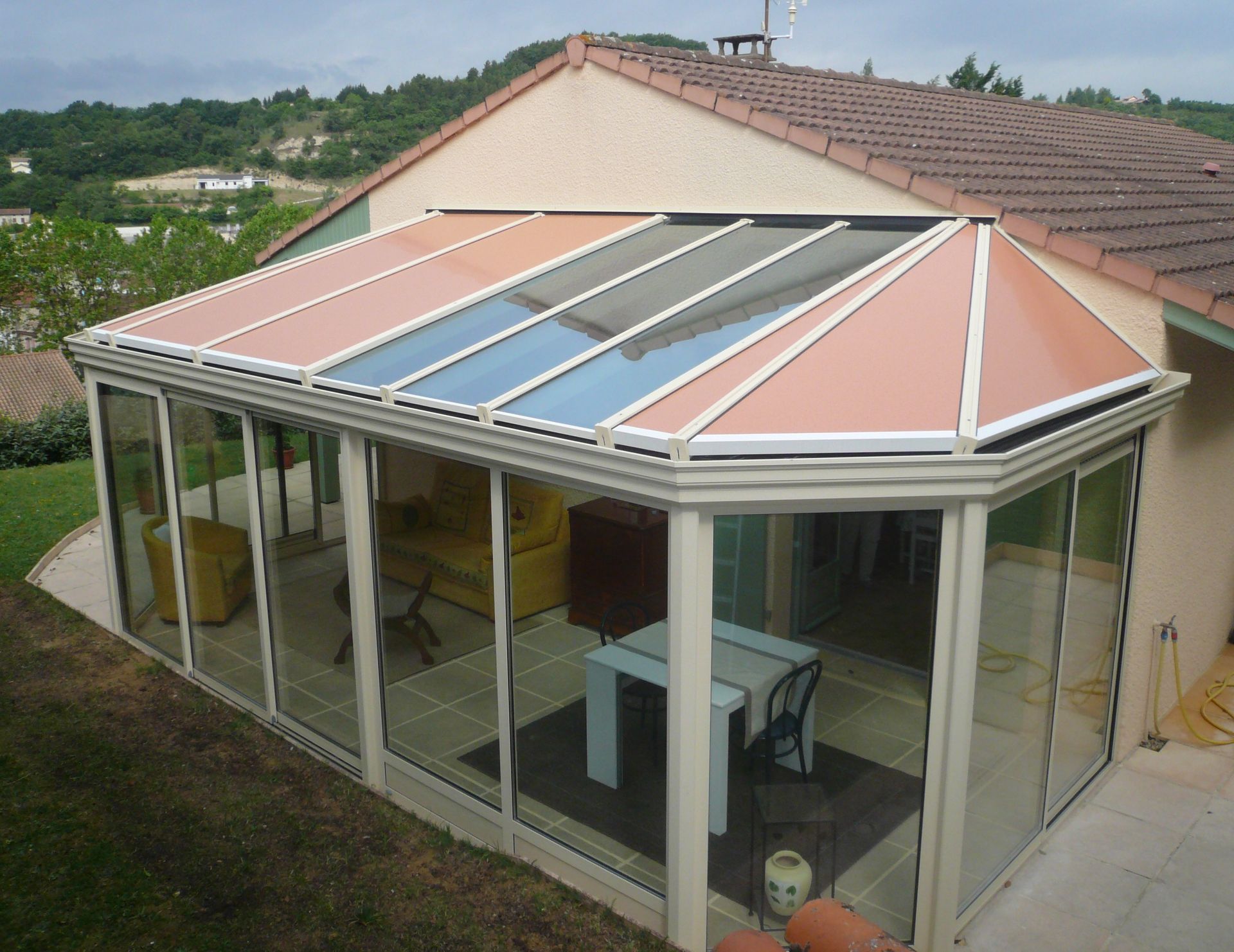 Une véranda beige attenante à une maison, dotée de grandes baies vitrées et d'un toit angulaire composé de parties pleines et vitrées.