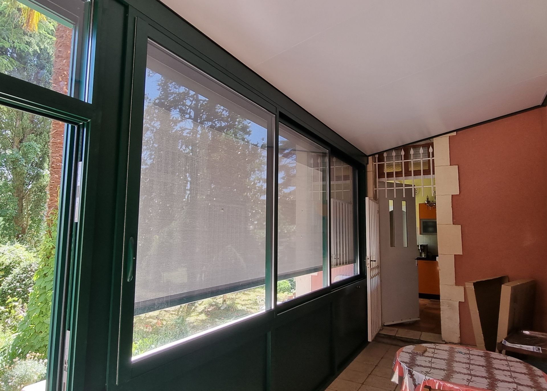 Une véranda avec des cadres de fenêtres vert foncé, des stores solaires foncés et une table, donnant sur une porte intérieure.
