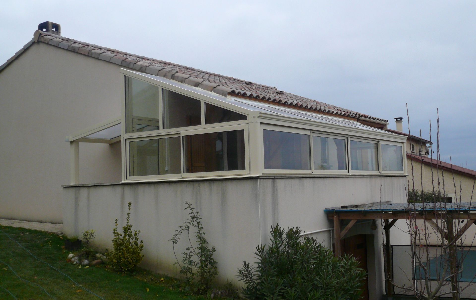 Une maison d'habitation couleur crème avec une véranda vitrée en pente, se détachant sur un ciel nuageux.