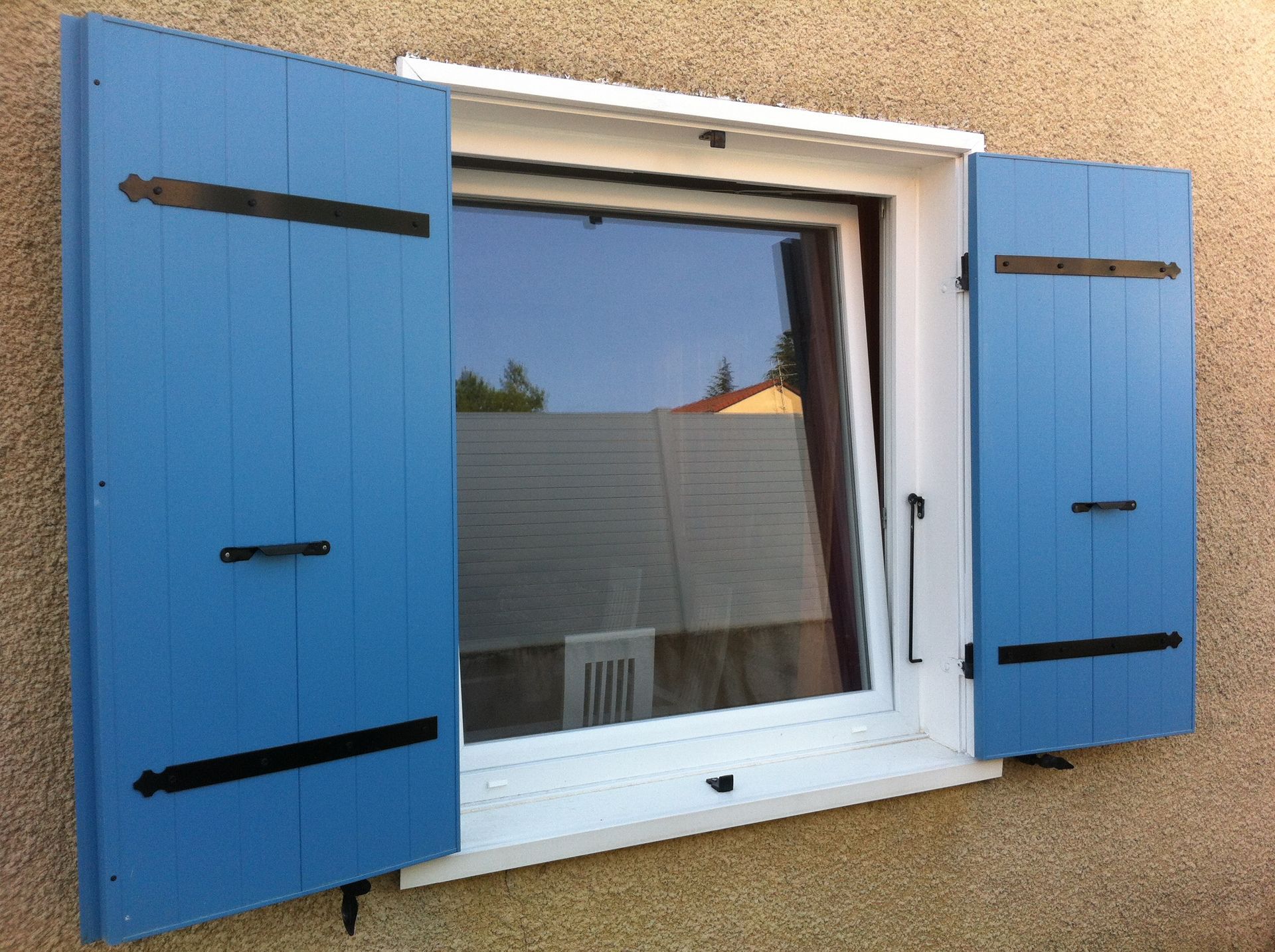 Une fenêtre carrée aux encadrements blancs et aux volets en bois bleu vif s'ouvre sur un mur extérieur beige texturé.