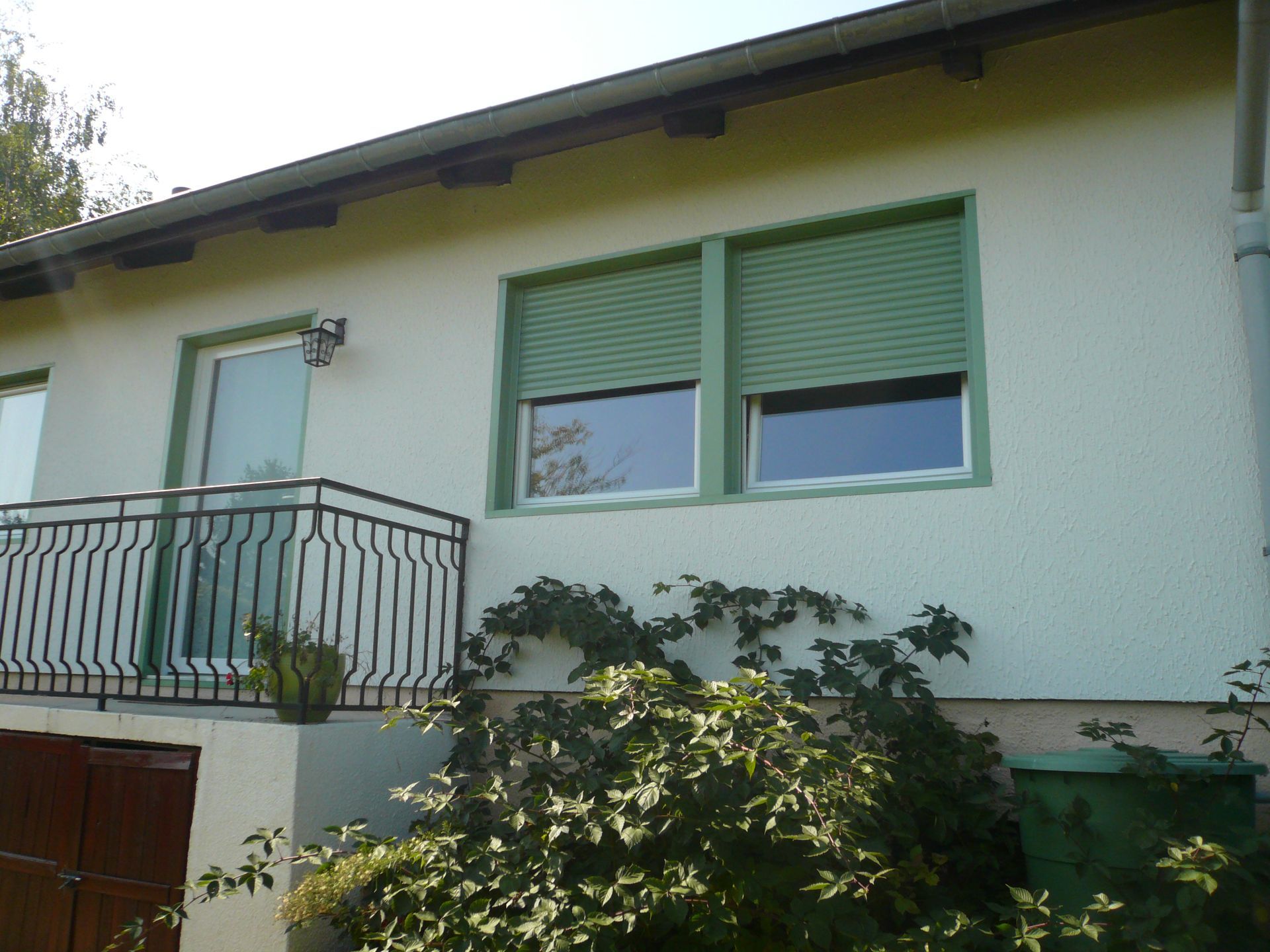 Vue extérieure d'une maison montrant un balcon avec une rambarde noire, une porte et deux fenêtres dont les stores verts sont partiellement baissés.