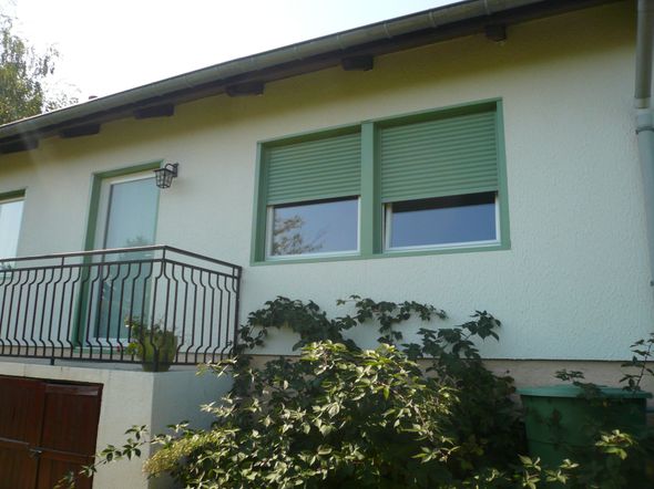 Vue extérieure d'une maison aux murs blancs, avec un balcon, des volets verts et un jardin en contrebas.