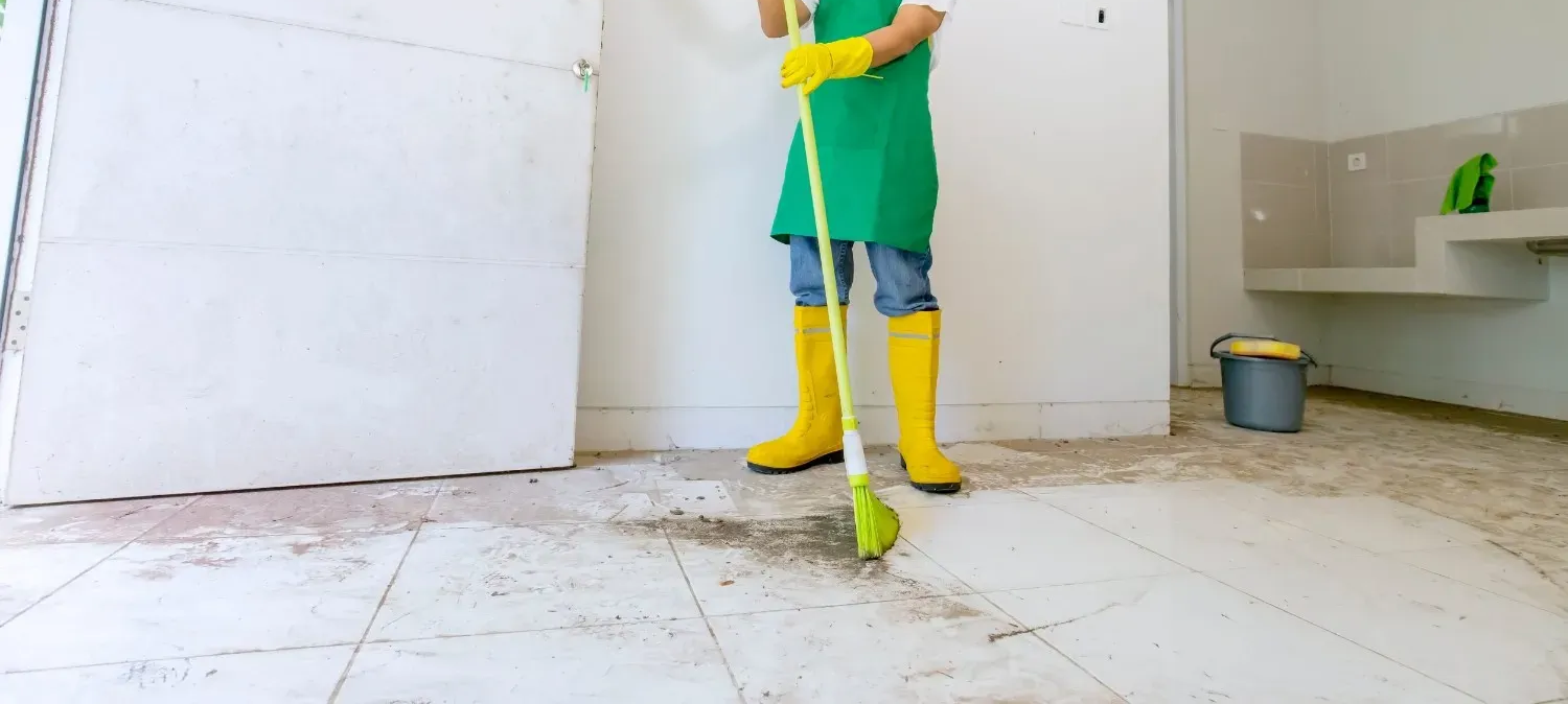 Persona con camisa verde y botas amarillas fregando el suelo de una habitación con baldosas.