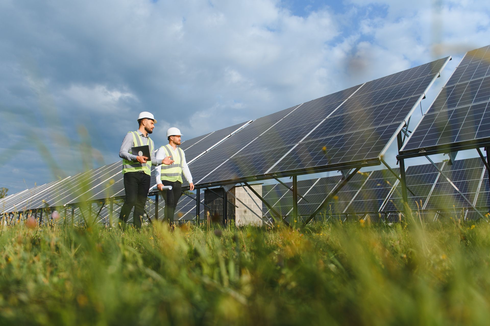 Deux ouvriers inspectent des panneaux solaires dans un champ herbeux sous un ciel nuageux.