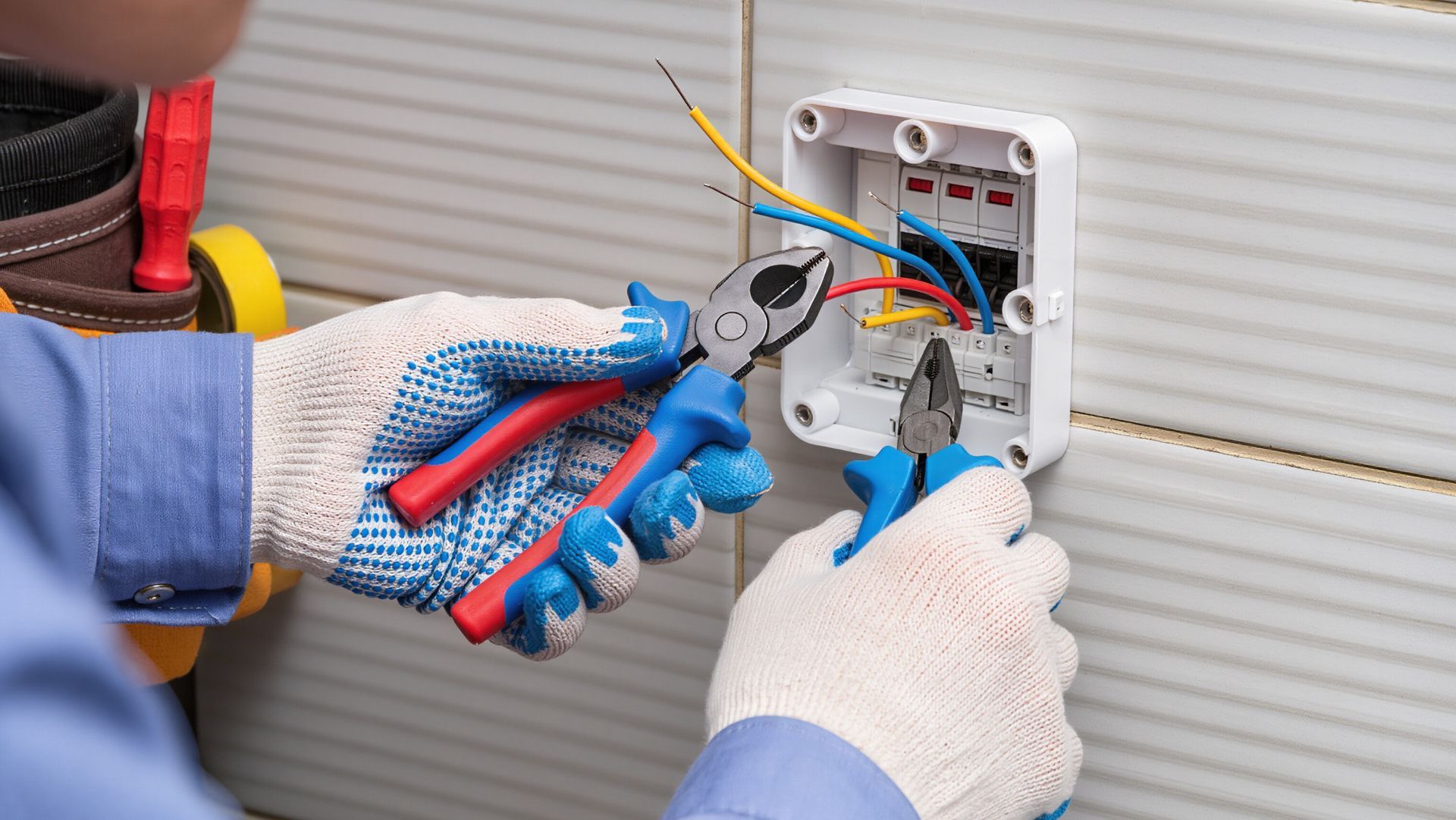 Un électricien ganté effectue le câblage d'une boîte de prise murale à l'aide d'outils et de câbles de couleur apparents.