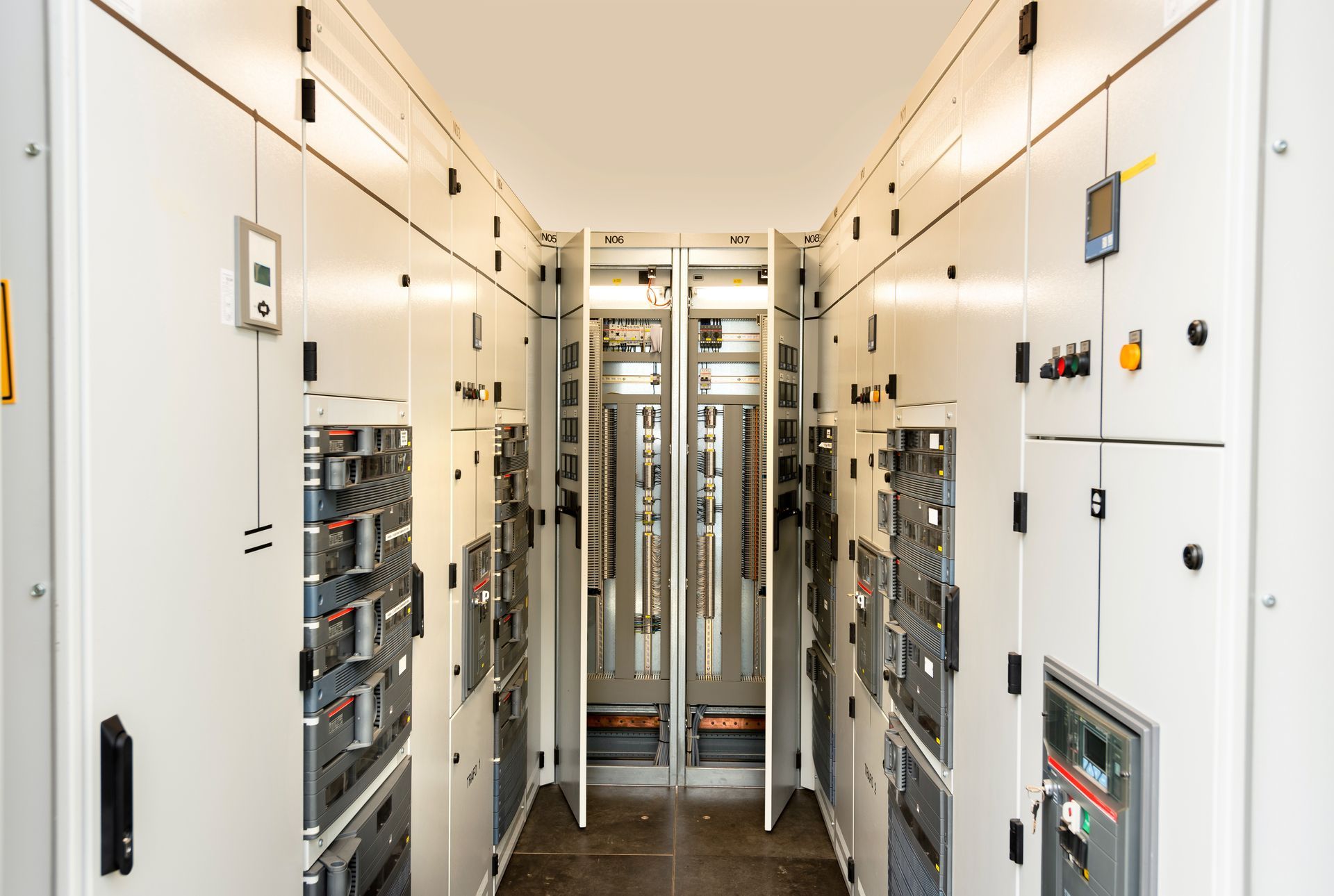 Salle de contrôle électrique étroite avec panneaux blancs, câbles et armoires électriques ouvertes de part et d'autre.