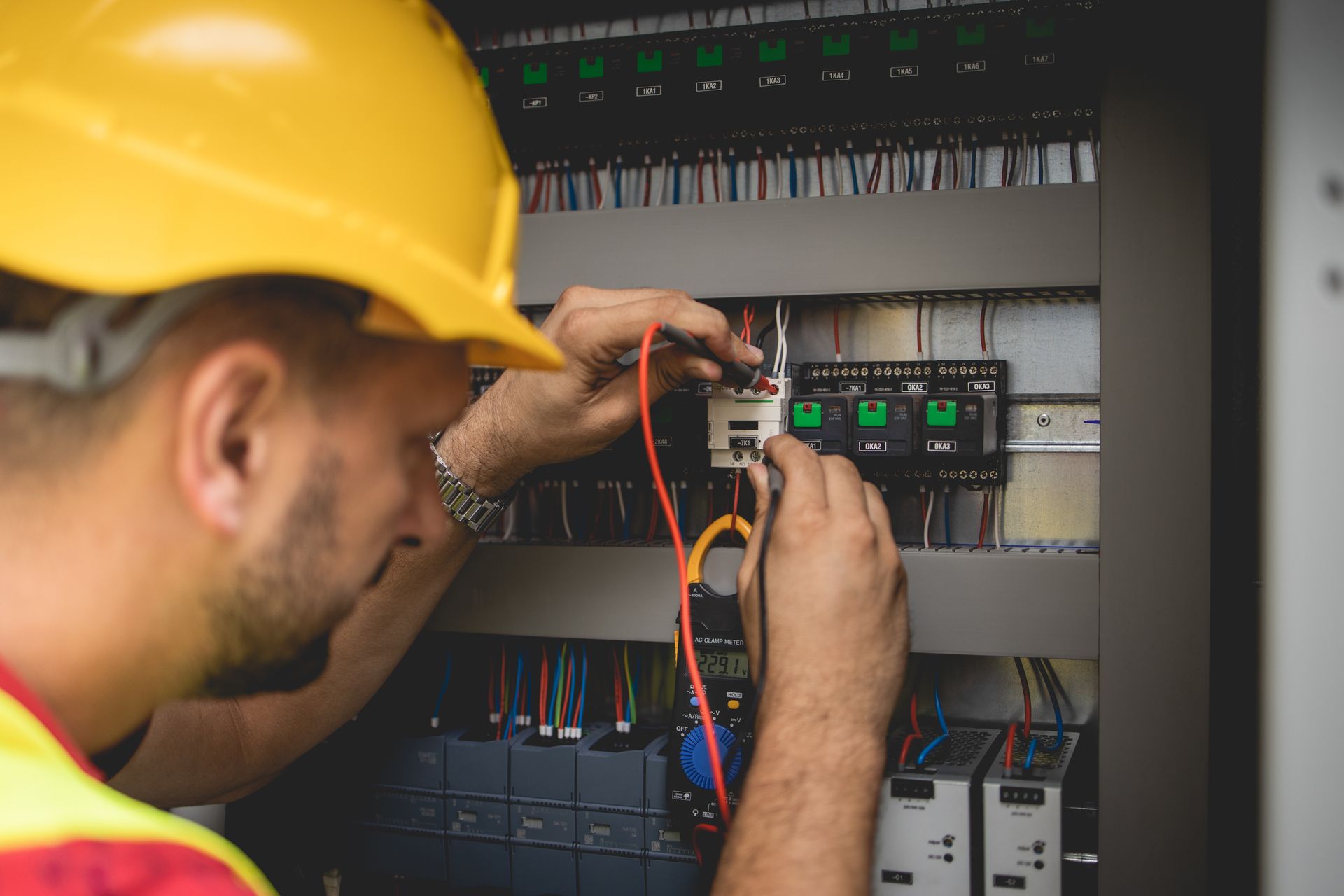 Un technicien inspecte le câblage électrique d'un panneau de commande, portant un casque jaune et un gilet de sécurité.