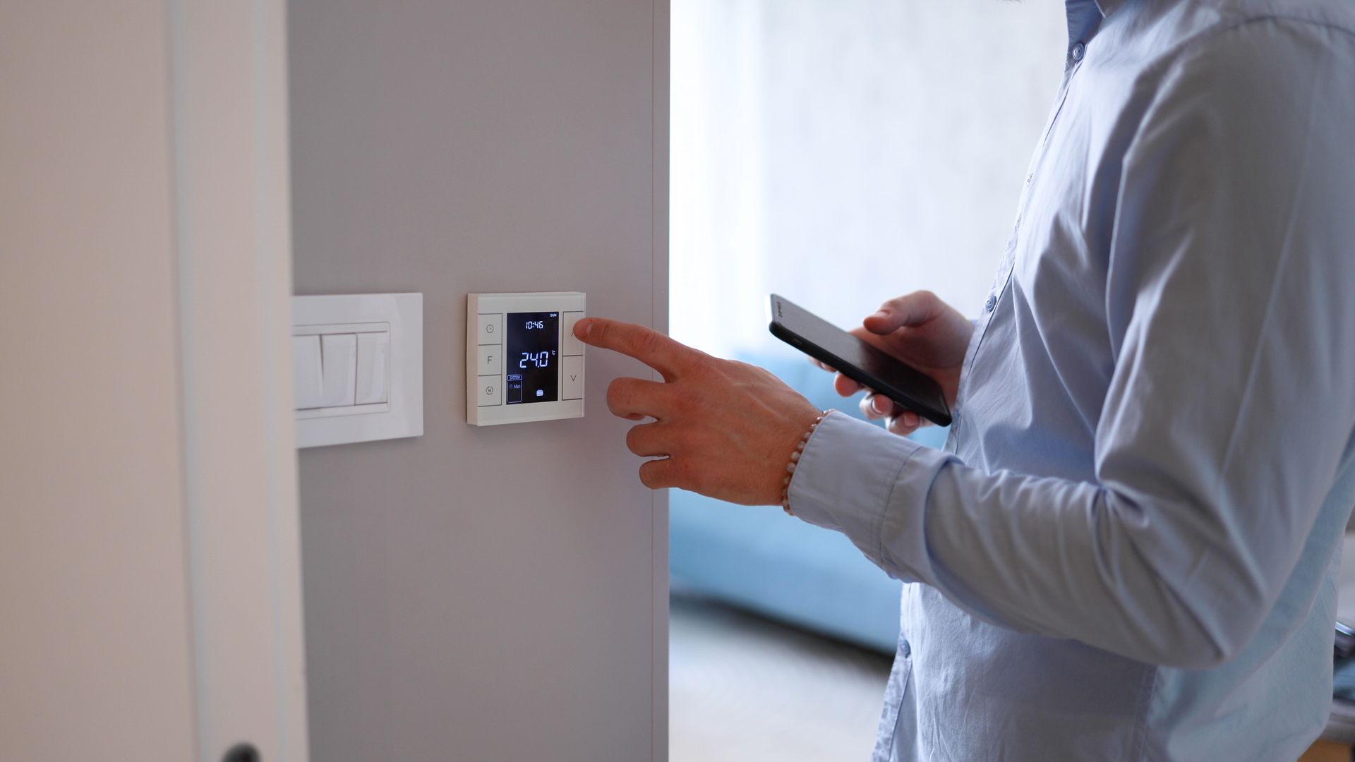 Une personne utilise un smartphone pour régler un thermostat mural dans une pièce lumineuse.