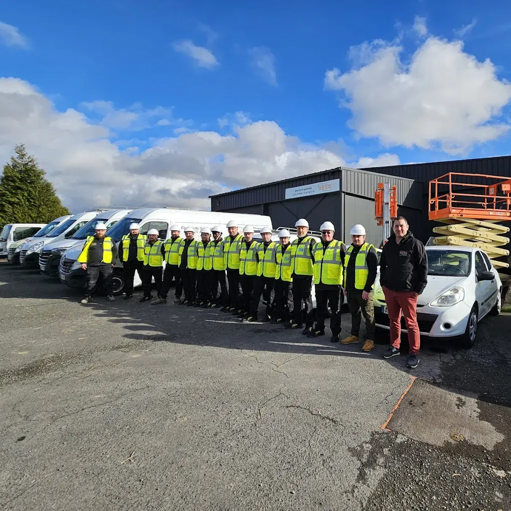 Des ouvriers vêtus de gilets à haute visibilité et de casques de chantier sont alignés à côté de camionnettes blanches devant un bâtiment industriel.