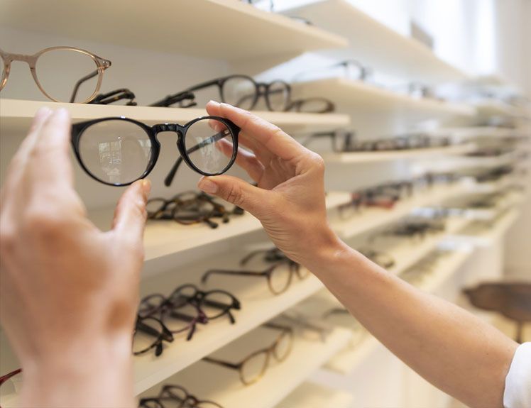 Des mains tenant des montures de lunettes noires dans une vitrine de magasin d’optique