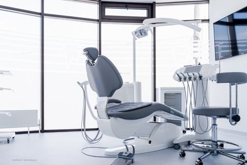 Helles Behandlungszimmer mit grauem Behandlungsstuhl, Arzthocker und medizinische Geräten für den Bereich Implantologie.