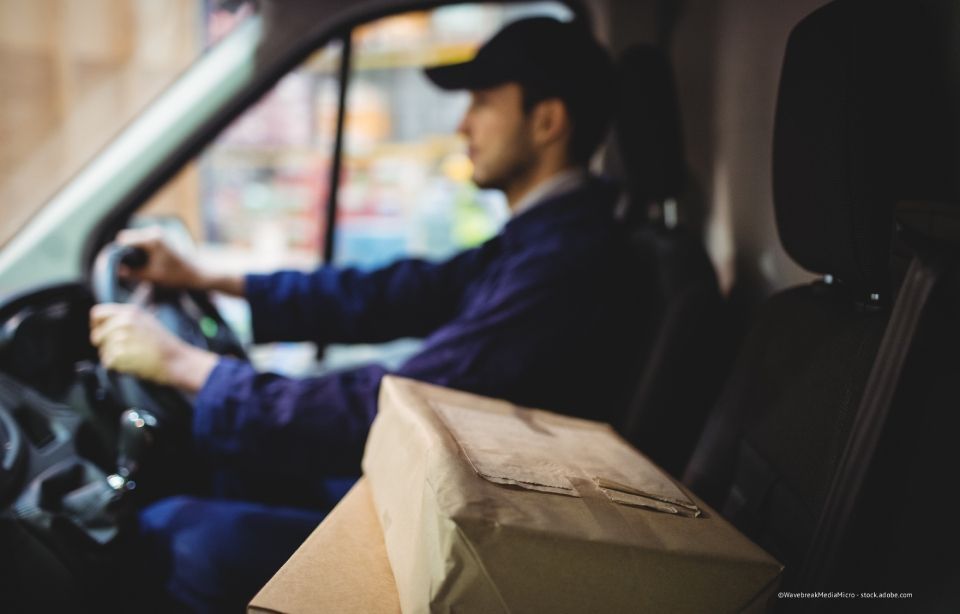 Lieferfahrer in dunkler Uniform am Steuer eines Lieferwagens, im Vordergrund ein Karton