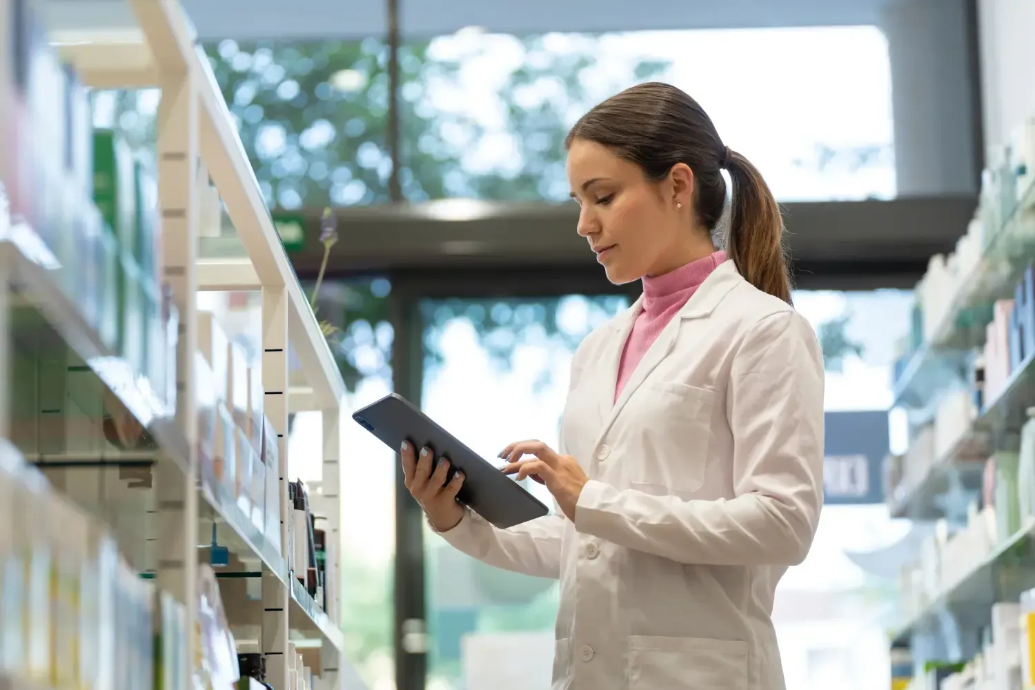 Apotheker im weißen Kittel benutzt ein Tablet in einem Apothekenregal.