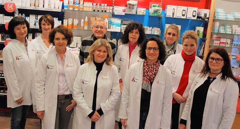 Eine Gruppe von Apothekern in weißen Kitteln steht in einem Apothekengang und lächelt in die Kamera.