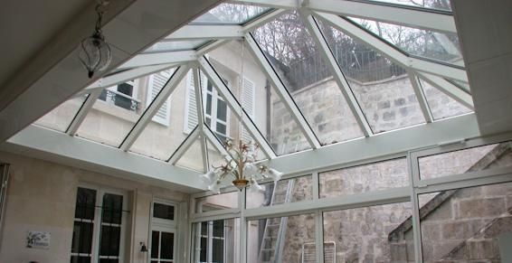 Véranda à toit de verre et cadres blancs, donnant sur une façade en pierre. Un lustre est suspendu à l'intérieur.