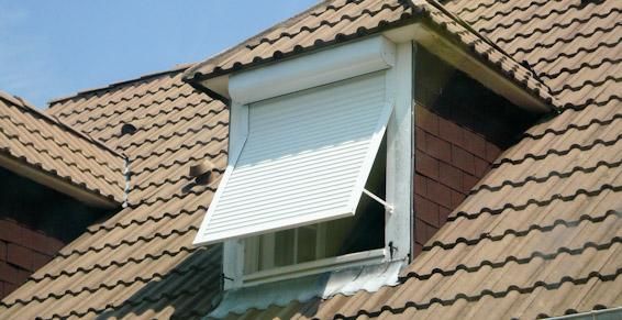 Volet blanc au-dessus d'une fenêtre sous un toit de tuiles brunes.