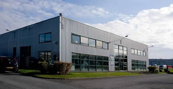 Bâtiment industriel gris avec fenêtres, situé sur une pelouse verte sous un ciel bleu nuageux.