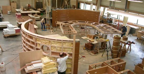 Atelier de menuiserie avec des ouvriers construisant une structure en bois incurvée ; outils et matériaux éparpillés alentour.