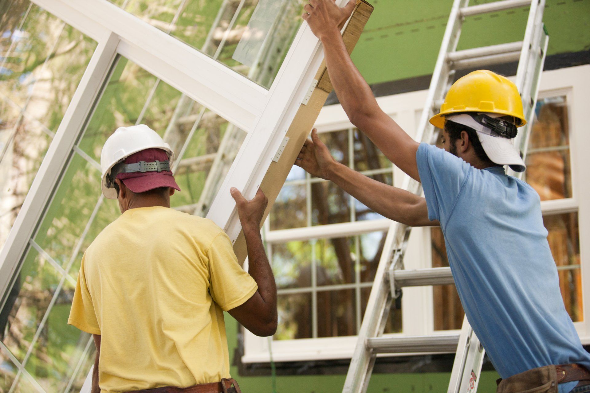 Deux ouvriers du bâtiment installent une fenêtre, l'un d'eux sur une échelle, à l'extérieur.