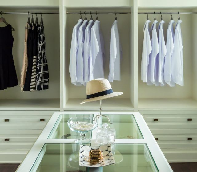 Dressing avec étagères blanches, robes et chemises blanches suspendues ; une table avec un chapeau et des objets décoratifs.