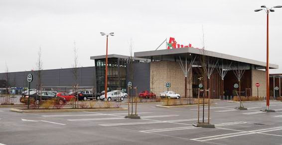 Façade d'un grand magasin avec parking ; ciel couvert. Bâtiment brun et gris, logo rouge au sommet.