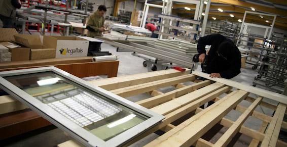 Un ouvrier assemble une fenêtre, entouré de matériaux et d'équipements. Une autre personne travaille à l'arrière-plan.