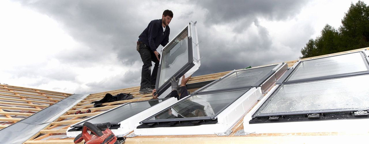 Un ouvrier installe un puits de lumière sur un toit en bois sous un ciel nuageux.