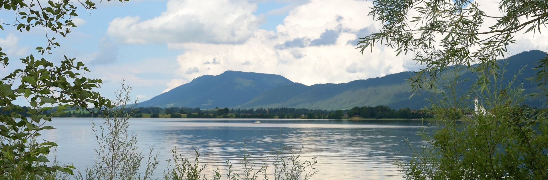 Bergsee mit ruhigem Wasser, bewaldetem Ufer und blauem Himmel mit Wolken.