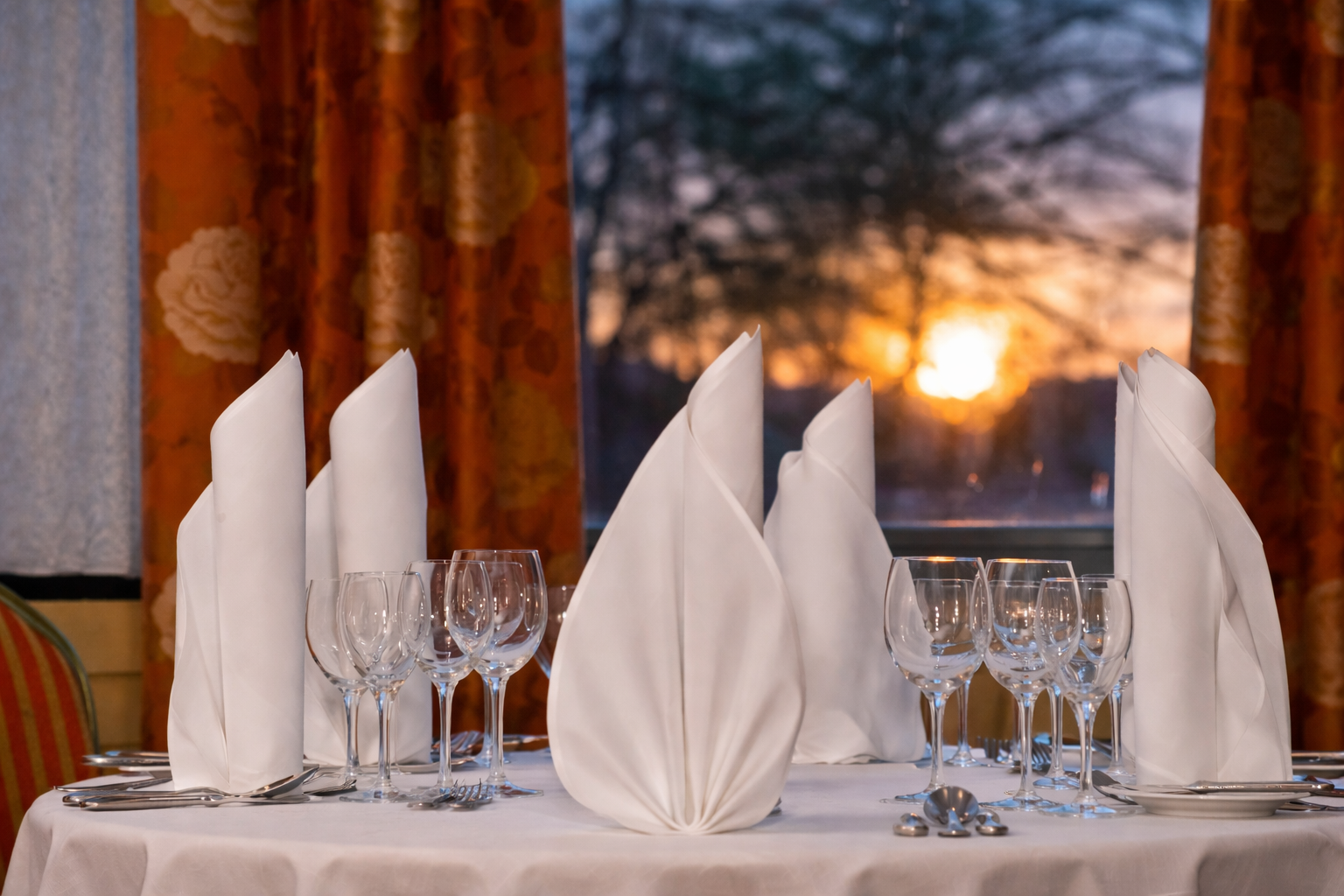 Elegant gedeckter Restauranttisch mit weißen Servietten, Gläsern und Blick auf den Sonnenuntergang durch ein Fenster