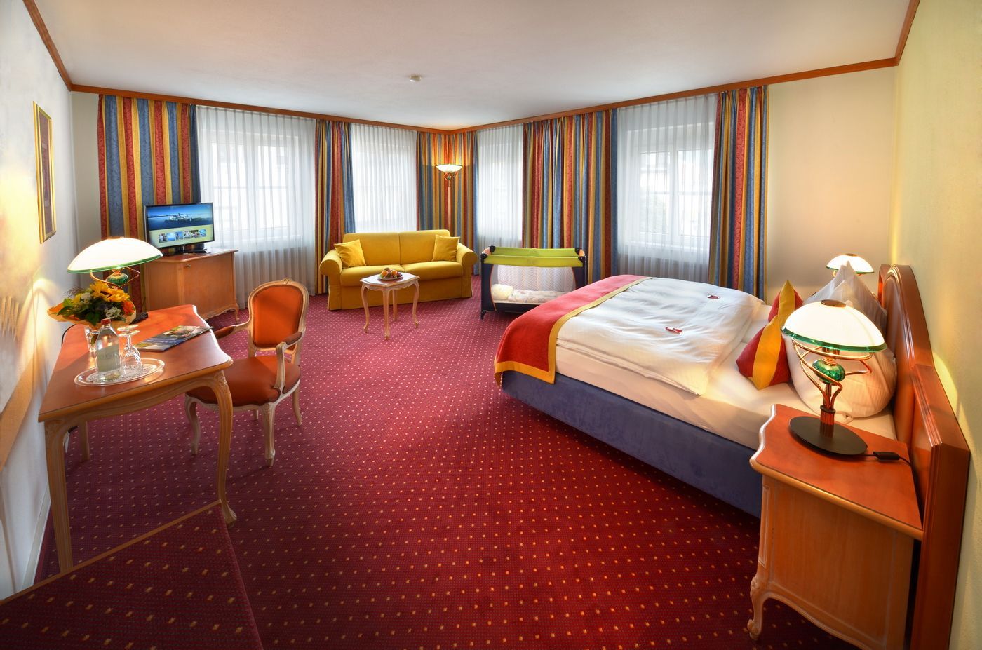 Hotelzimmer mit zwei Betten, rotem Teppich, Schreibtisch, Sofa und großen Fenstern mit Vorhängen.