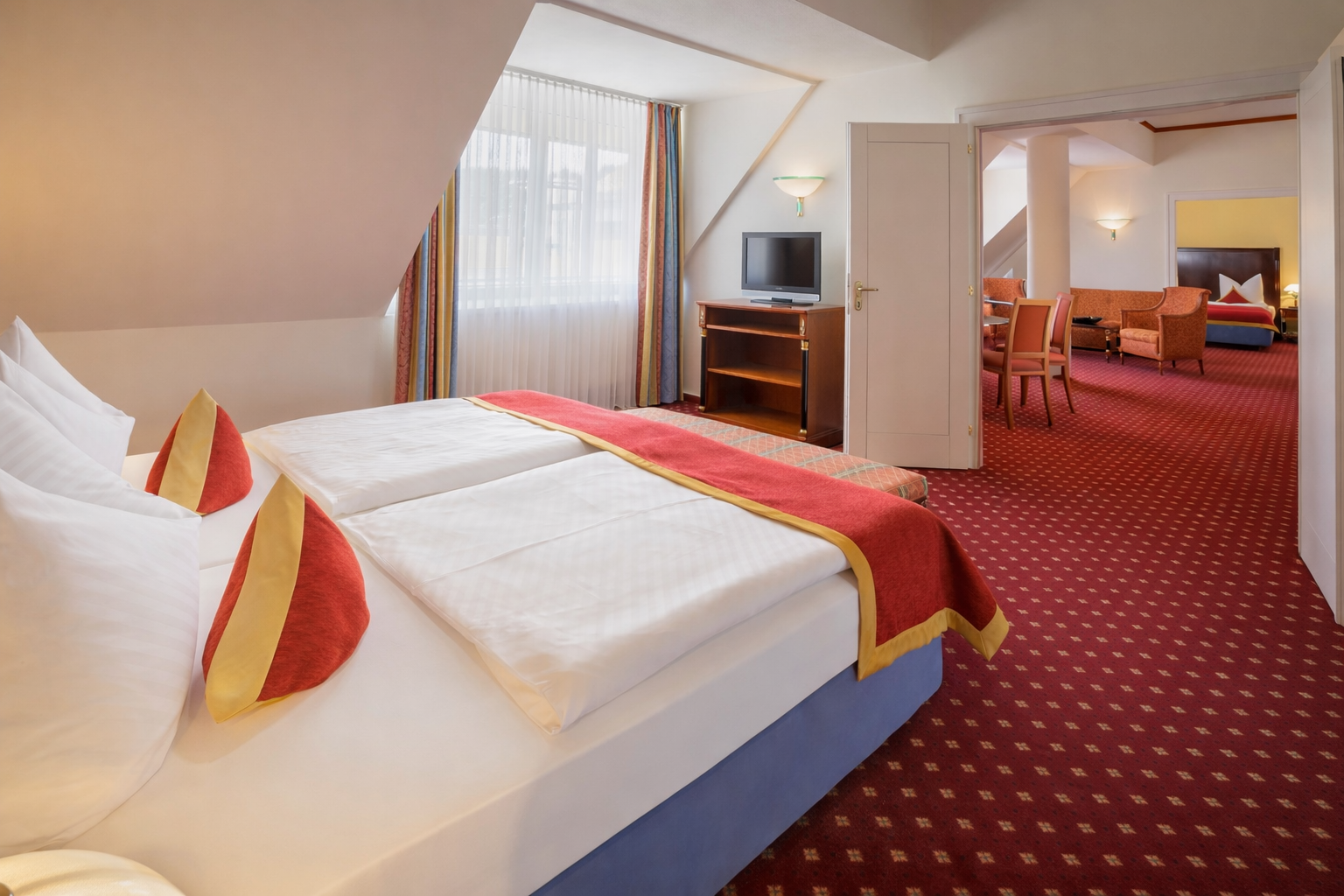 Hotelzimmer mit zwei Einzelbetten, rotem Teppich und freiem Blick durch einen Torbogen in einen Sitzbereich